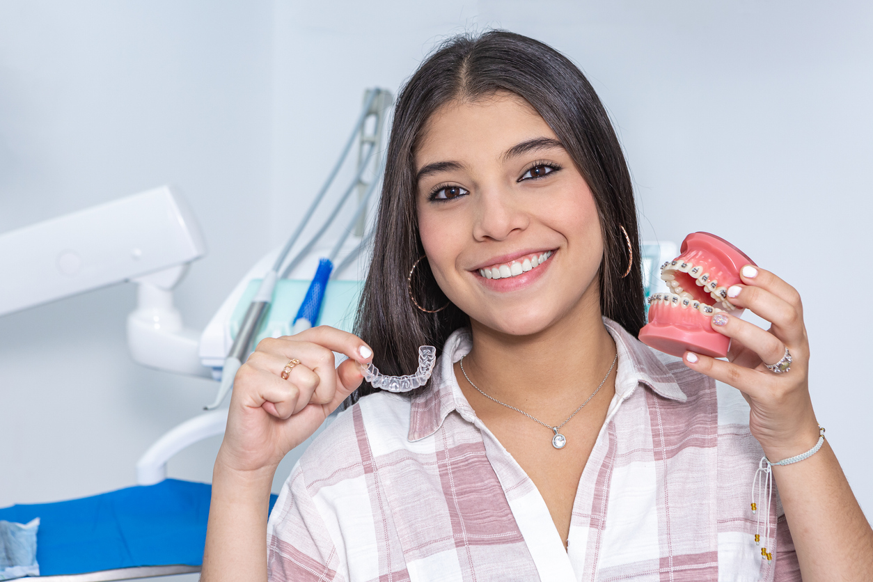 Delighted Hispanic female teenaged with long dark hair in casual clothes smiling and looking at camera while holding teeth model with braces and transparent retainer in each hand at dental clinic. The image represents how to determine which orthodontic option is best.